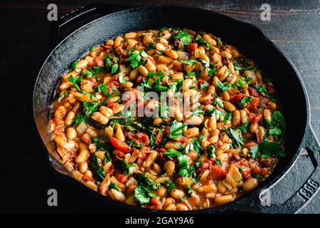Vegan Spanish Beans with Tomatoes in a Cast Iron Skillet: Cannellini beans, diced tomatoes, and spinach garnished with parsley in a cast-iron pan Stock Photo
