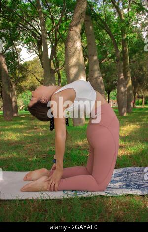 Vertical image of an unrecognizable young woman doing a camel pose outside on a green grass in a sunny day on summer. Pilates Outdoor 2021. Stock Photo