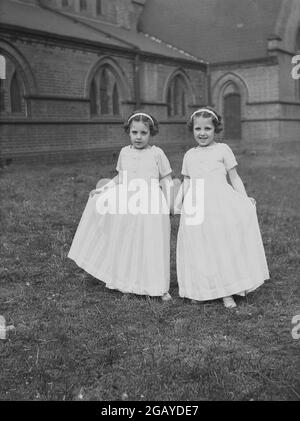 1956, historical, two young girls, sisters, holding hands as they stand for a photo outside in the church grounds, in their outfits for the special May Day celebration and carnival, Leeds, England, UK. Stock Photo