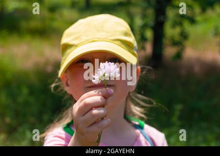 Positive emotional young lady smiles, looks on wild flower that holds in hand. Cheerful pretty child girl in yellow baseball cap, pink shirt, stands o Stock Photo