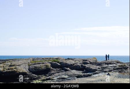 Trearddur Bay, Anglesy, Wales. Stony shoreline, holiday makers standing on low cliffs, looking out to sea. Seaside landscape. Blue sky and copy space. Stock Photo