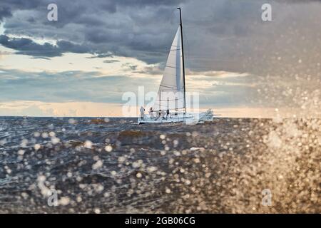 The lonely sailboat on the horizon through splashes of sea at sunset, the storm sky of different colors, big waves, sail regatta, cloudy weather, only Stock Photo
