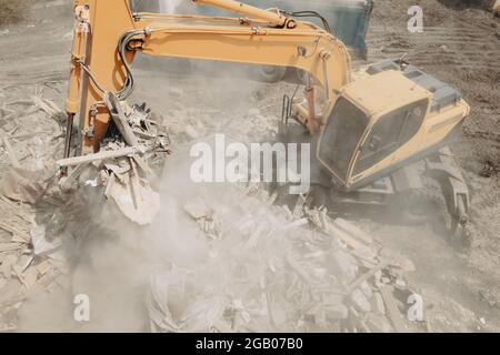 Excavator breaks building and loads construction waste into truck with its bucket. Demolition of building. Stock Photo