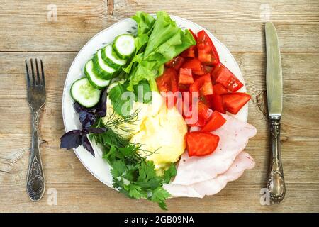 Scrambled eggs with tomato, cucumber, herbs, ham and lettuce on old wooden table, fork and knife Stock Photo