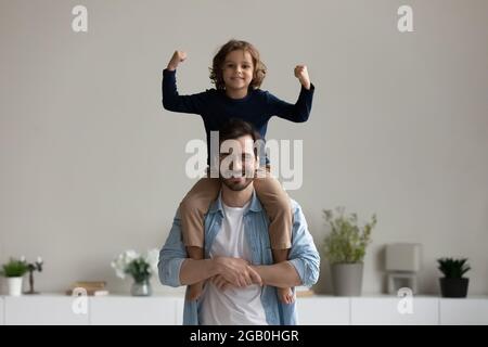 Excited boy sitting on dads neck, making strength hand gesture Stock Photo