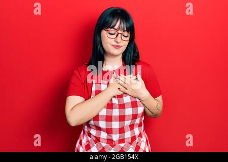 Young hispanic woman wearing cook apron and glasses smiling with hands on chest with closed eyes and grateful gesture on face. health concept. Stock Photo