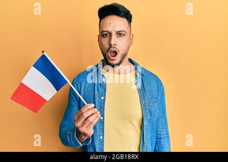 Young arab man holding france flag scared and amazed with open mouth for surprise, disbelief face Stock Photo