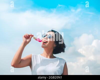 Smiling Asian woman short hair in casual white sleeveless shirt wearing sunglasses holding pink popsicle on blue sky background in summertime. Women e Stock Photo