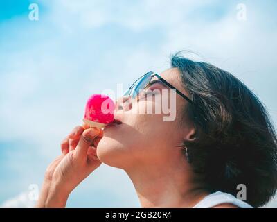 Smiling Asian woman short hair in casual white sleeveless shirt wearing sunglasses holding pink popsicle on blue sky background in summertime. Women e Stock Photo