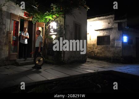 (210802) -- HEFEI, Aug. 2, 2021 (Xinhua) -- Night watchman Lu Lei talks with a villager while 'dageng' in Xidi Village, Yixian County of Huangshan City, east China's Anhui Province, July 29, 2021. It is a serene night as the luminous moon gleams in the sky. With a gong and mallet in hand, Lu Lei sets out to perform 'dageng'. Lu, a post-80s man, has been a night watchman as a substitute for his father, a member of Xidi's 'dageng' team, in the ancient village loved by many for its picturesque scenery. Lu works at a tourism company in the county seat of Yixian during the day and returns home Stock Photo