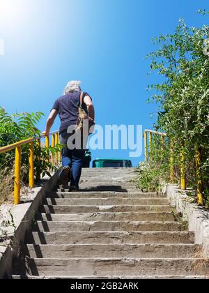 The gray-haired man climbs the concrete stairs in bright sunlight Stock Photo