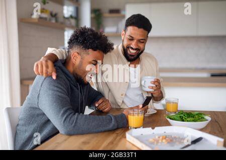 Portrait of young adult brothers in kitchen indoors at home, using smartphone when eating pizza. Stock Photo