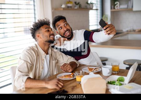 Portrait of young adult brothers in kitchen indoors at home, taking selfie. Stock Photo