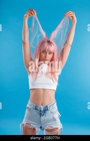 Funny female model in casual wear playing with pink hair, holding strands of it up over her head, standing isolated over blue studio background Stock Photo