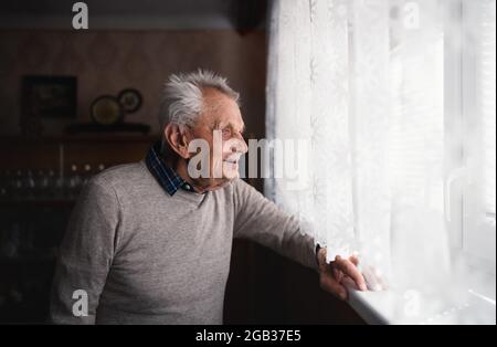 Portrait of elderly man standing indoors at home, looking out through window. Stock Photo