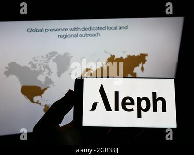 Person holding mobile phone with logo of American digital media company Aleph Holding on screen in front of web page. Focus on phone display. Stock Photo