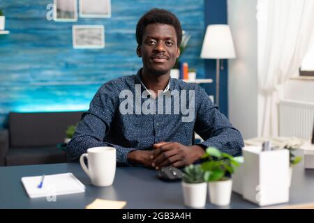 African american man freelancer smiling to camera, young person in modern flat apartment working from home using computer. Black businessman doing work tasks Stock Photo