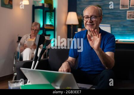 Retired old couple with handicap sitting indoors having walking support supplies. Senior man with crutches using laptop computer video call technology and elder woman disabled in wheelchair Stock Photo