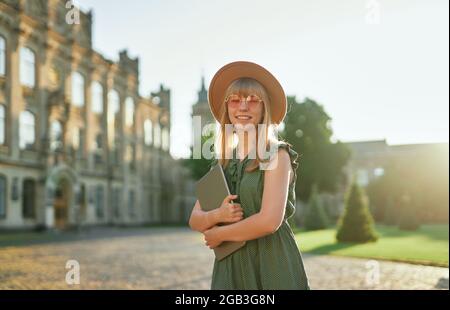 Studying abroad, student portrait or education concept. Cute cheerful blonde female university or college student with laptop wearing green dress and hat standing in campus. High quality image Stock Photo