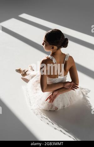 Side view of ballerina in white dress and pointe shoes sitting in sunlight on grey background Stock Photo