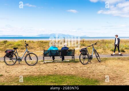 Two elderly cyclists sitting on a bench with their cycles beside them as a young man on a skateboard runs by, overlooking the Firth of Clyde towards t Stock Photo