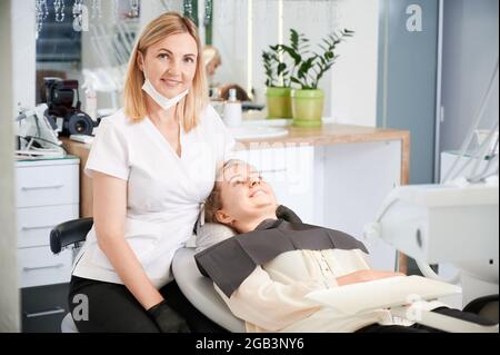 Horizontal snapshot of pretty, young female dentist, wearing uniform, rubber gloves, smiling, sitting at her working place in well equipped dental office. Female patient during dentist's visit Stock Photo
