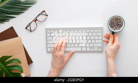 Woman typing on computer keyboard and drinking coffee at workplace. Flat lay office desktop workspace with female hands on white background Stock Photo
