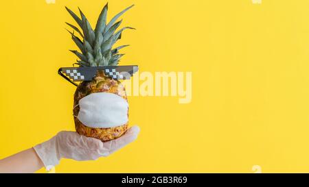 Funny pineapple face in a protective medical mask. Doctor hand in rubber glove holds summer tropical fruit pineapple in sunglasses. Safe travel Stock Photo