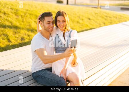 Cheerful young couple hugging and making video call on phone sitting relaxed on bench in city park on bench in summer sunny day. Stock Photo