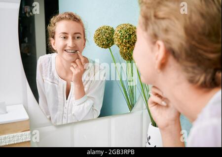 Woman with orthodontic brackets on teeth looking in the mirror. Charming woman in white elegant shirt checking braces on teeth in bathroom. Concept of Stock Photo