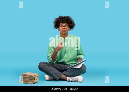 Thoughtful black guy student in glasses taking notes, holding notepad and pen, sitting over blue studio background Stock Photo
