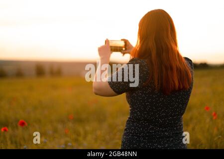 Back view of a young girl in a summer dress makes a photo on her smartphone in an autumn field at the golden hour Stock Photo