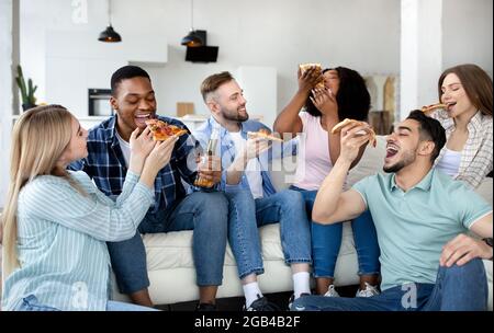 Positive diverse friends having home party, sitting on sofa, eating pizza with beer, having fun together Stock Photo