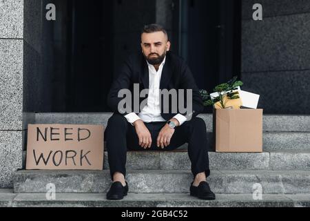 Portrait of fired mature businessman with paper poster saying need work and box of personal things, sitting on stairs Stock Photo
