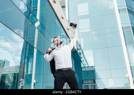 Ecstatic mature businessman celebrating success and raising hand up, standing outdoors near office center, empty space Stock Photo
