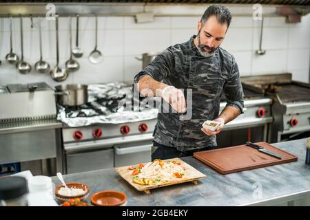 Professional cook is preparing meal in restaurant's kitchen. He is inserting spices in meal after cooking. Stock Photo