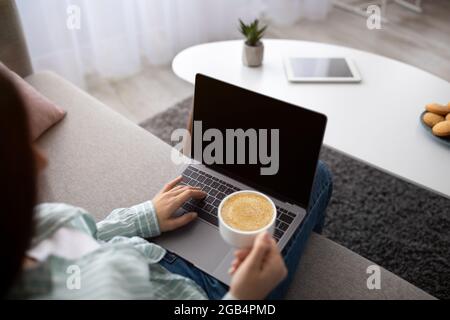 Young Indian woman with coffee sitting on sofa and using laptop computer with blank screen at home, mockup Stock Photo