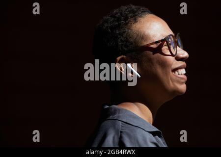 Close-up of African young woman in eyeglasses listening to music in wireless headphones and smiling isolated on black background Stock Photo