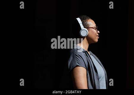 Side view of African young woman in wireless headphones listening to music and smiling against the black background Stock Photo