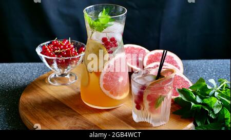 Grapefruit lemonade with mint, lemon, currants and ice in a decanter and a glass with black plastic tubes on a background of fruit. Cooling, refreshing, fruit, summer drink. Horizontal close up Stock Photo