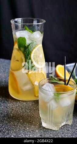 Lemon lemonade with mint, lemon and ice in a decanter and a glass with black plastic tubes on a background of fruit. Cooling, refreshing, fruit, summer drink. Vertical close up Stock Photo