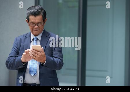 Serious senior entrepreneur answering text messages or checking mobile application on smartphone when standing outdoors Stock Photo