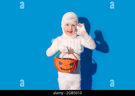 Child in mummy costume holding basket of chocolates in front of blue background. Stock Photo