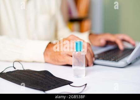 Focus on sanitizer, Unrecognizable man working on laptop at home while medical face mask and sanitizer at working desk - concept of coronavirus or Stock Photo