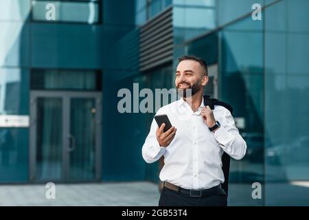 Handsome mature businessman browsing net on cellphone during coffee break, walking near office, copy space Stock Photo