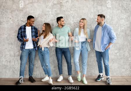 Group of diverse young friends having conversation, chatting with each other against grey studio wall, full length Stock Photo