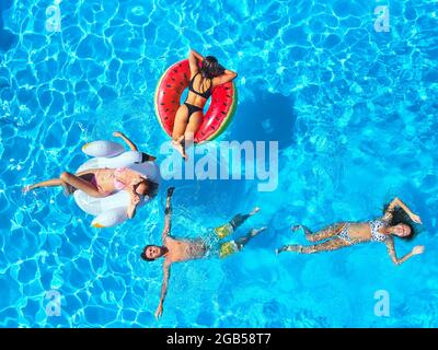 Aerial of friends having party in swimming pool with inflatable flamingo, swan, mattresses. Happy young people relax at luxury resort on sunny day. Vi Stock Photo