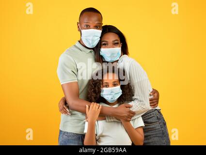 Young african american parents and their daughter wearing protective medical masks, embracing over yellow background Stock Photo