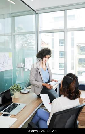 Serious frowning female entrepreneur leaning on desk and talking to her colleague planning work, analyzing financial documents and graphs Stock Photo