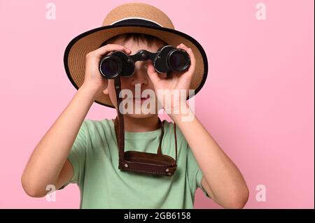 Close-up portrait of boy in summer straw hat looking through old vintage binoculars against pink background with copy space. Travel, tourism, summer, Stock Photo